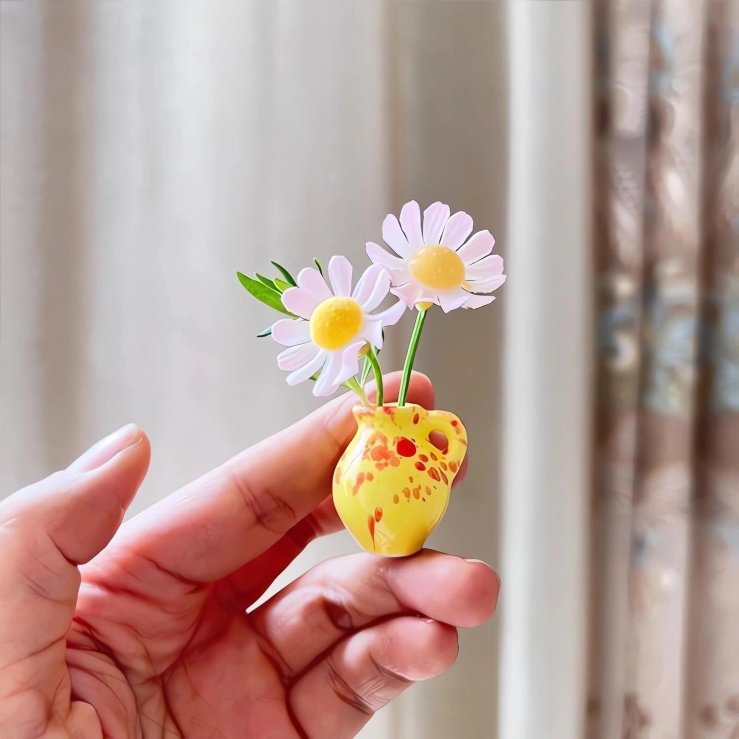 Hand holding a small yellow vase with two pink flowers against a neutral background