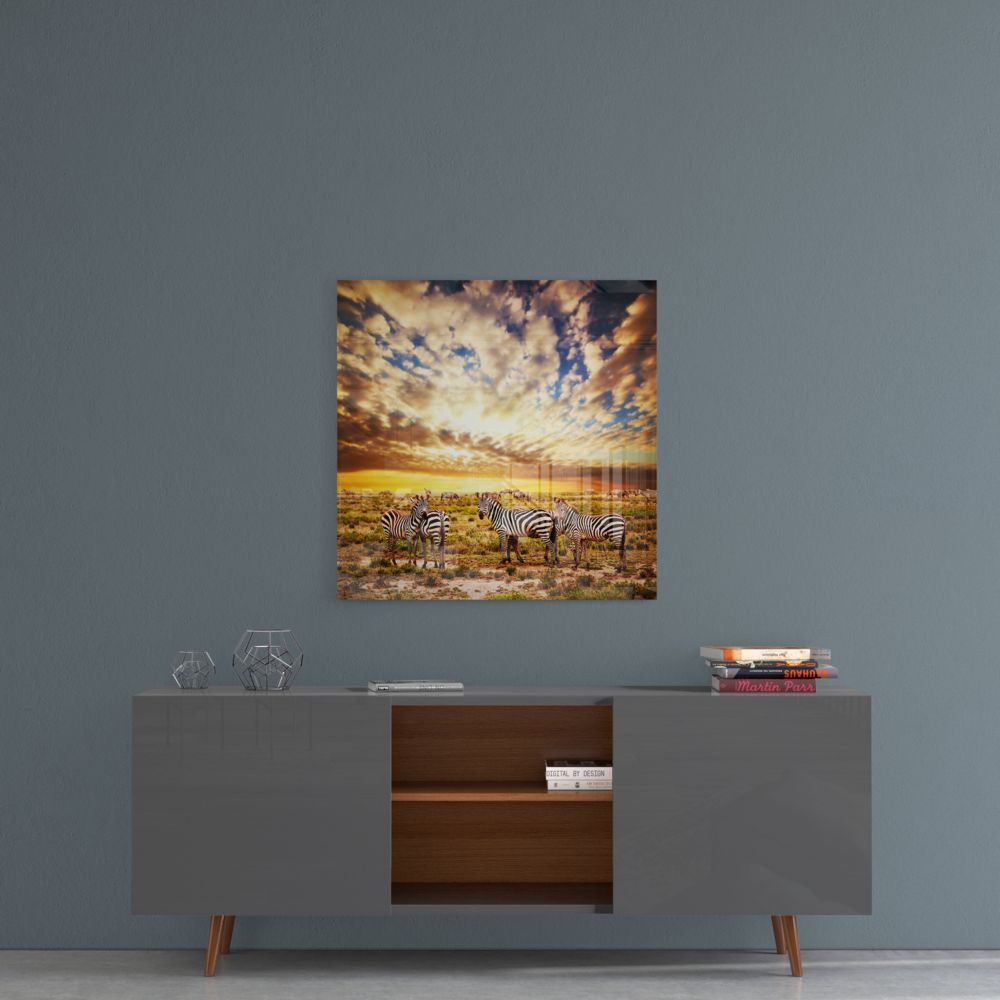 Zebra herd at sunset glass wall art on a blue-gray wall above a gray sideboard with wood shelves
