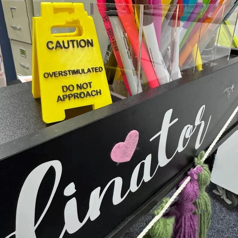 Mini desk sign with “Caution Overstimulated Do Not Approach” displayed beside office pens.