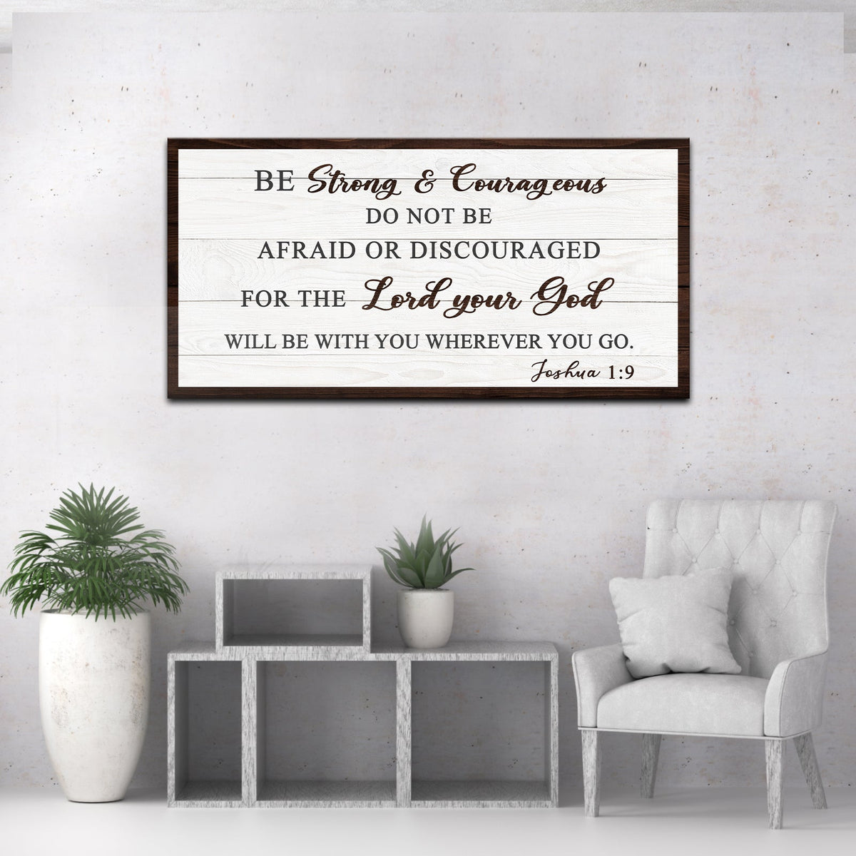 Joshua 1:9 Be Strong and Courageous canvas sign in the dark-border style, on a gray wall beside an armchair