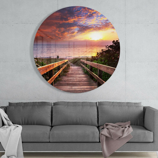 Beach boardwalk sunset round glass wall art above a gray sofa with draped throws on a pale concrete wall