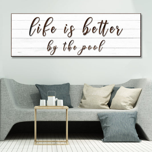Life Is Better Poolside Script canvas sign in white with brown script, hung above a gray sofa with pillows