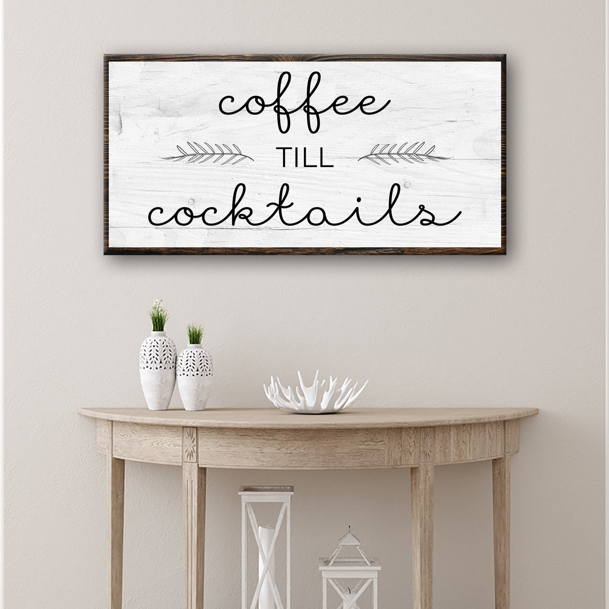 White canvas sign reading coffee till cocktails in black script with leaf sprigs, above a pale wood console table