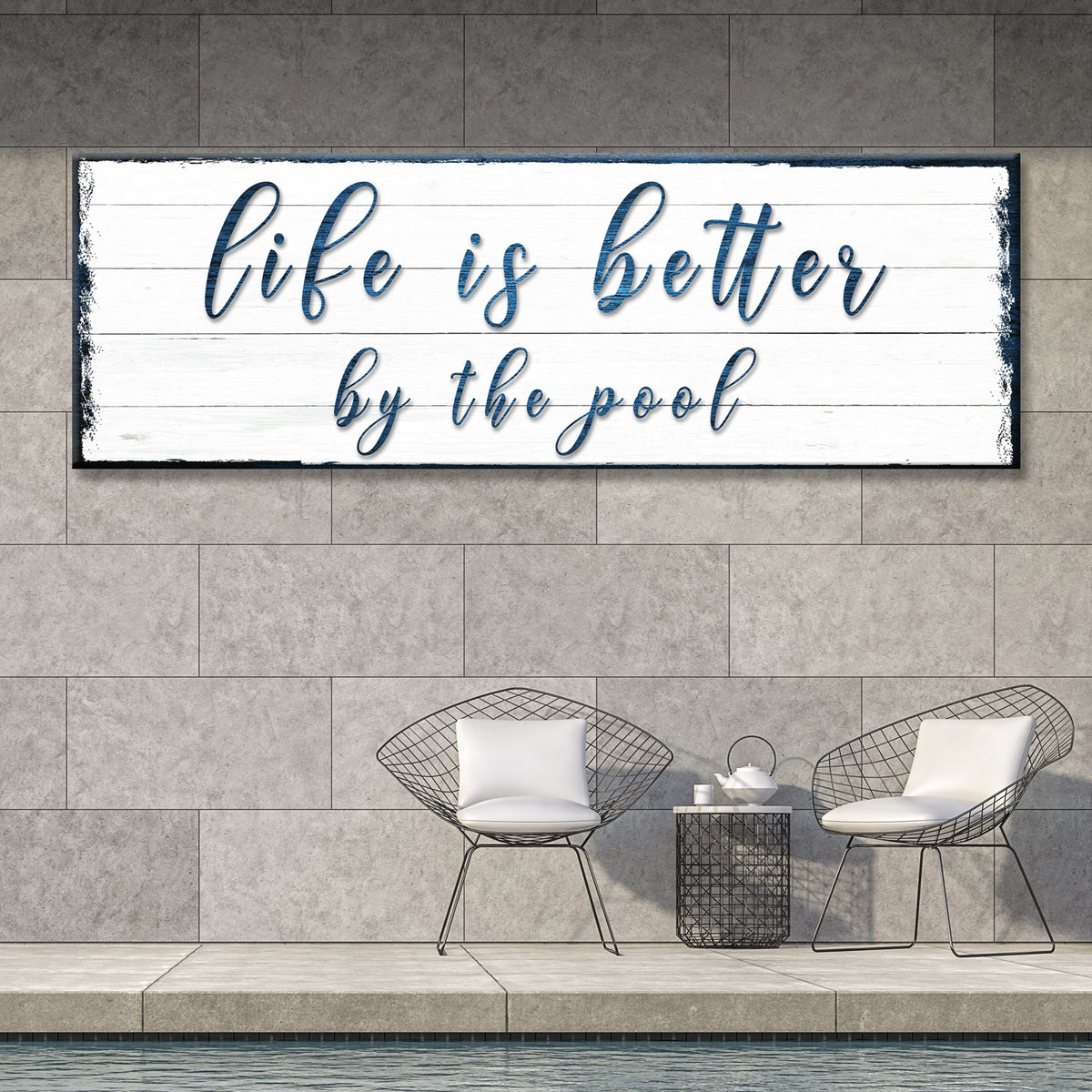 White plank-look style of the poolside script canvas sign with blue lettering on a gray stone wall above two wire chairs