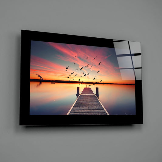 Birds over a wooden pier at pink sunset glass wall art with a black border hung on a plain gray wall