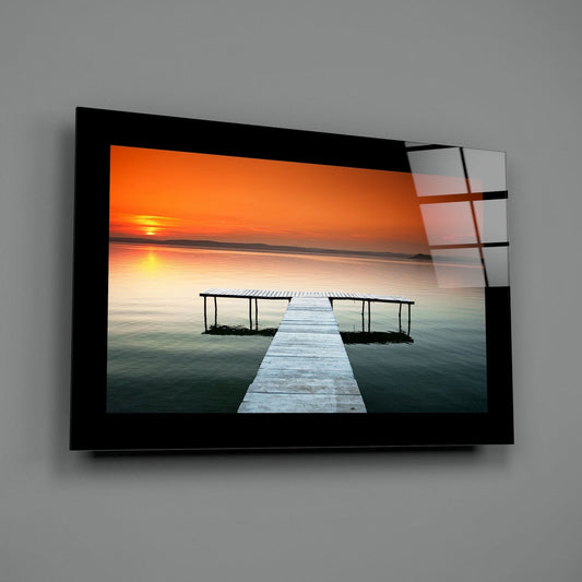 White pier on a teal lake at orange sunset glass wall art with a black border hung on a plain gray wall