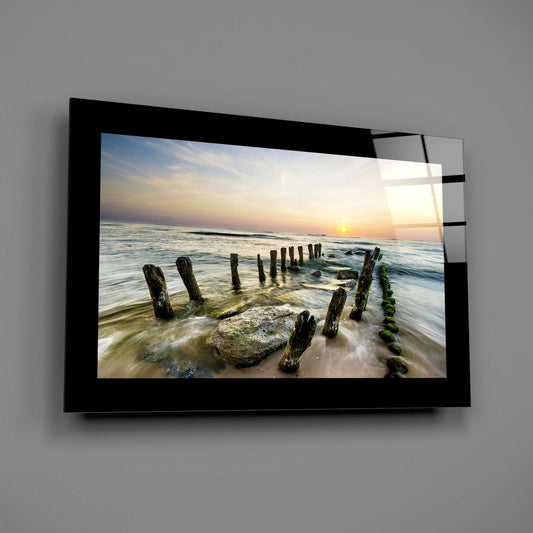 Mossy breakwater posts at a beach sunrise glass wall art with a black border hung on a plain gray wall