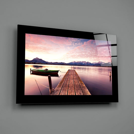 Green rowboat by a mountain lake pier glass wall art with a black border hung on a plain gray wall