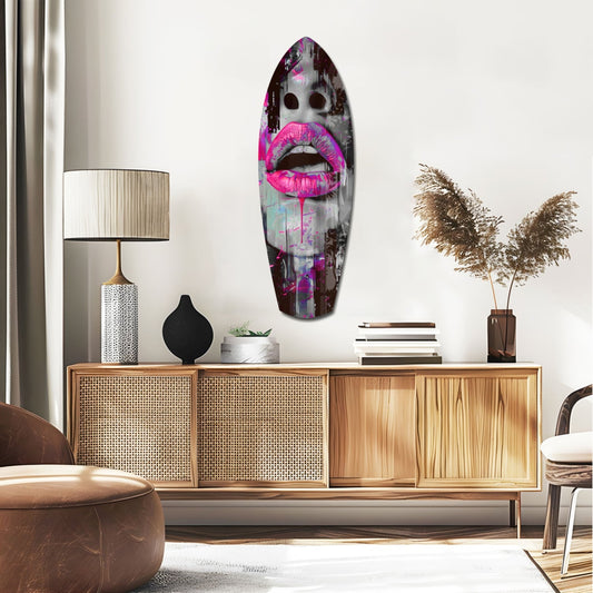 Pink drip lips on gray face surfboard glass wall art above a rattan sideboard with a table lamp and pampas grass