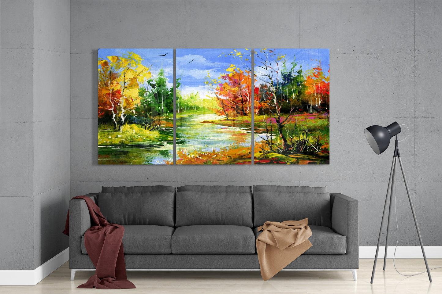 Autumn river landscape 3-panel glass wall art set above a gray sofa with red and tan throws beside a tripod lamp