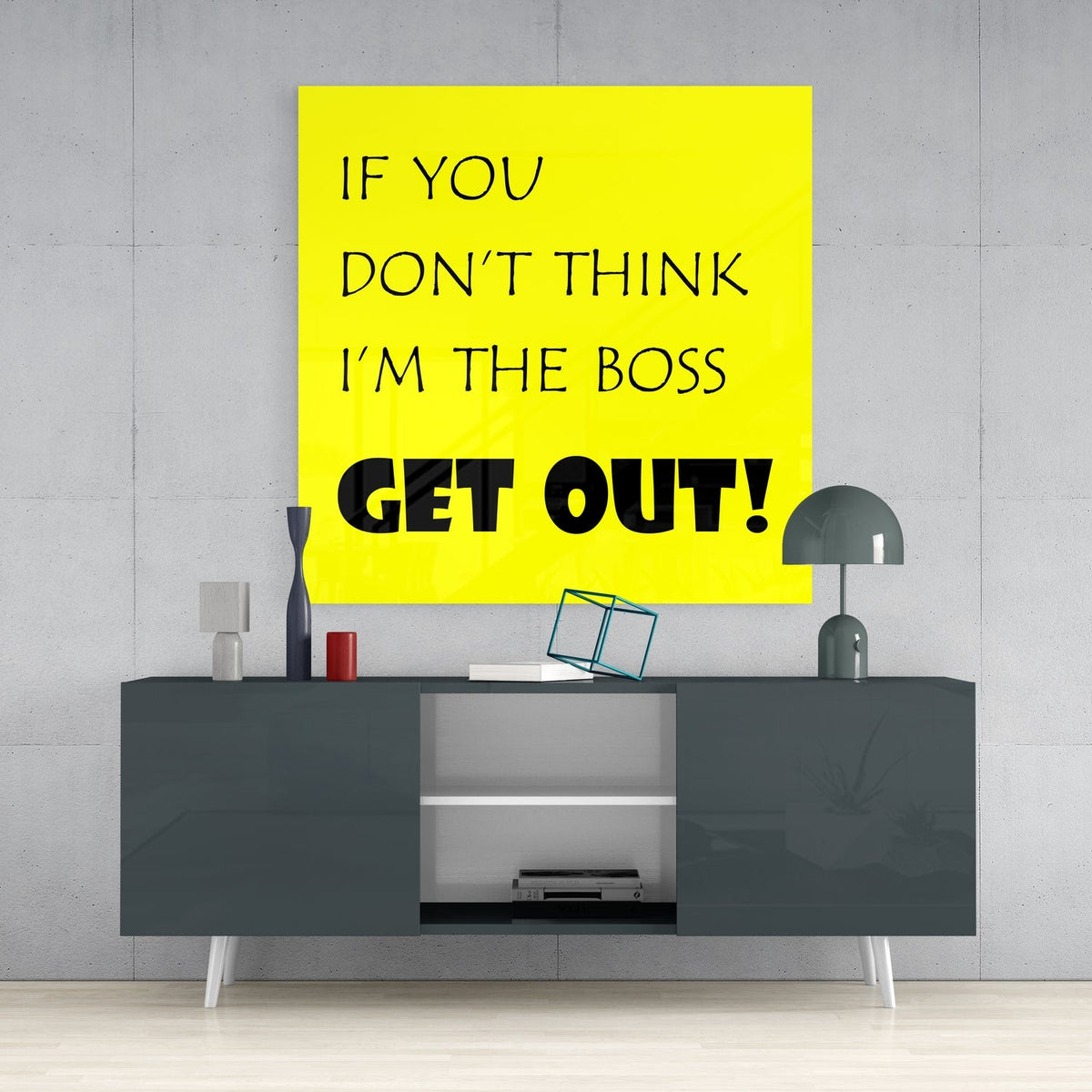 Funny boss quote in black text on yellow square glass wall art above a dark gray sideboard with a gray table lamp