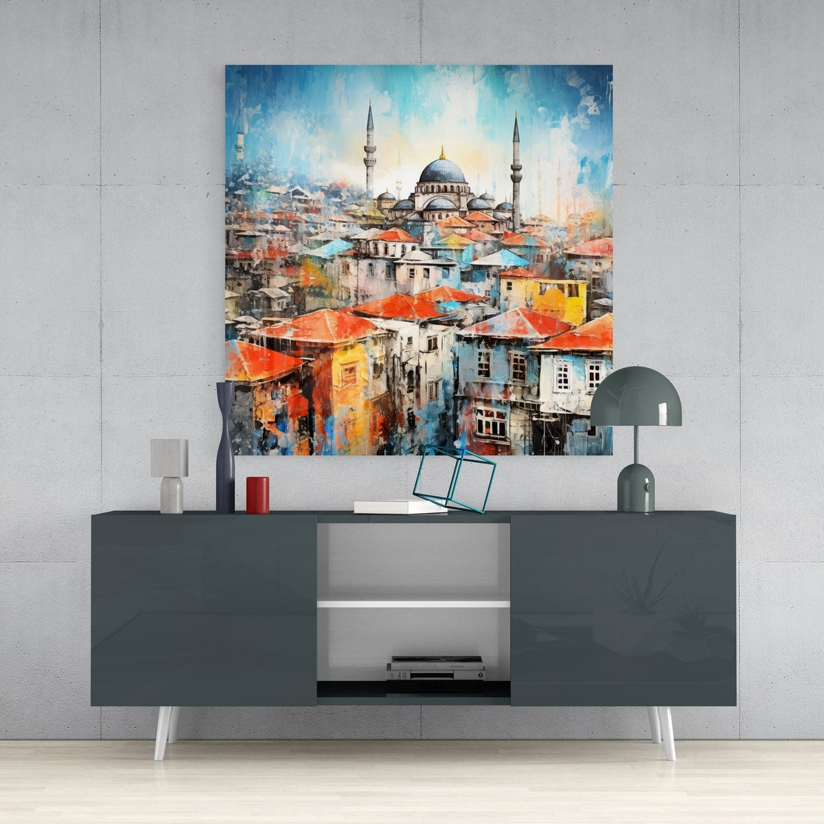 Hillside town and mosque glass wall art above a dark gray sideboard with a small lamp