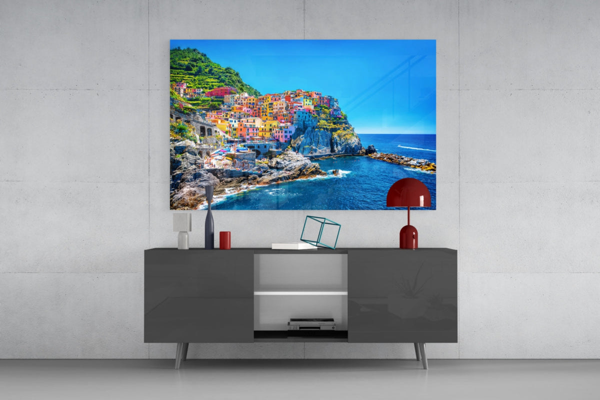Manarola village glass wall art above a gray sideboard with a red table lamp