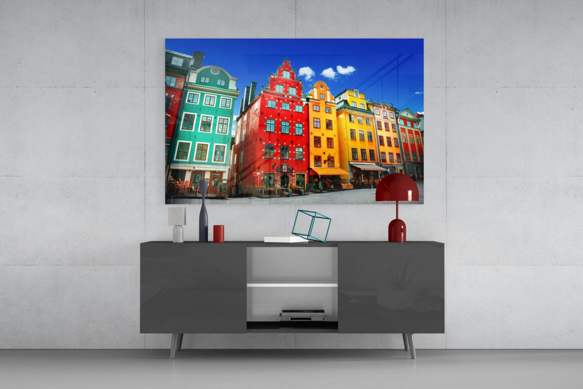 Stockholm Old Town houses glass wall art above a gray sideboard with a red table lamp