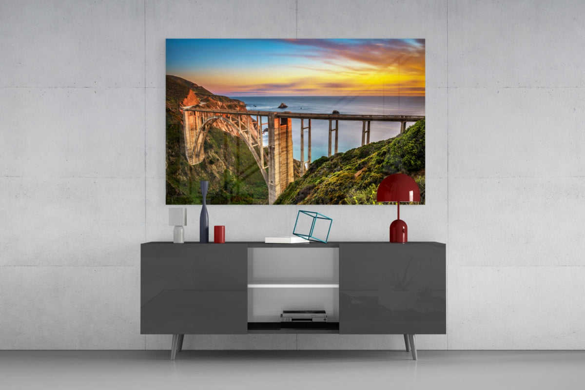 Bixby Creek Bridge glass wall art above a gray sideboard with a red lamp on a pale concrete wall