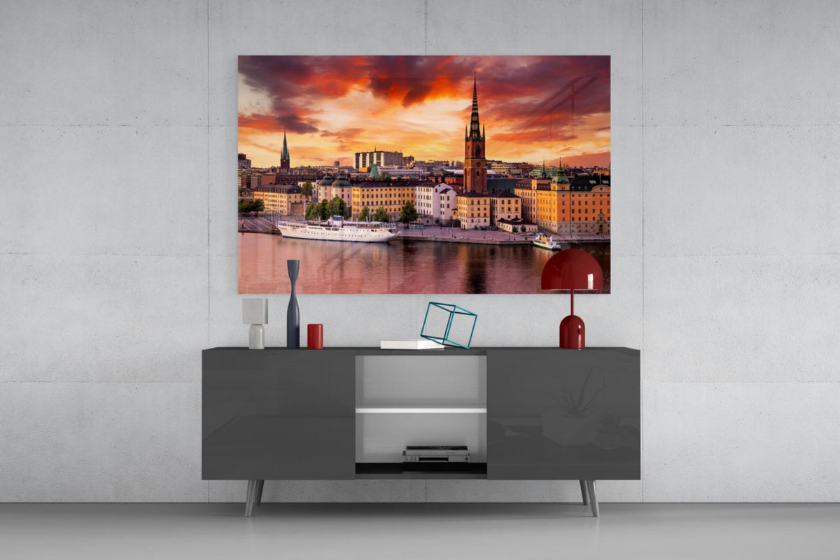 Stockholm waterfront at sunset glass wall art above a gray sideboard with a red table lamp