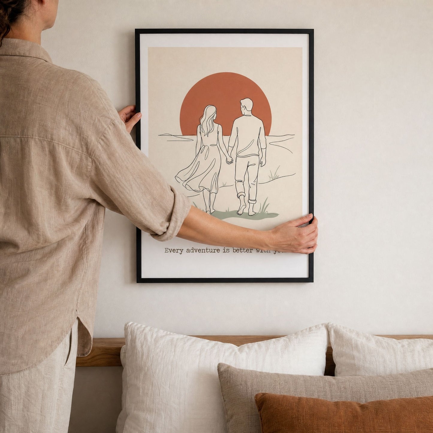 A hand steadying the black framed couples print while levelling it on the wall above a bed.