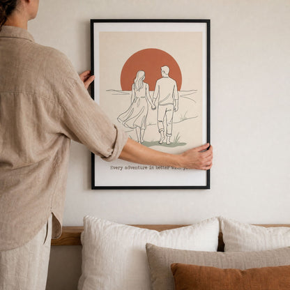 A hand steadying the black framed couples print while levelling it on the wall above a bed.