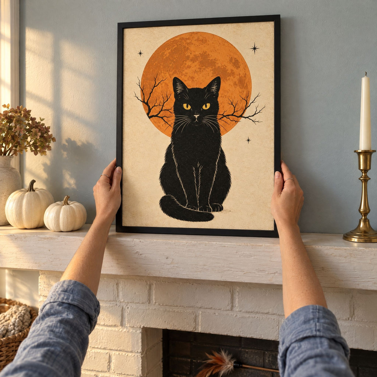Black cat and full moon print in a black frame being set on an October mantel beside white pumpkins