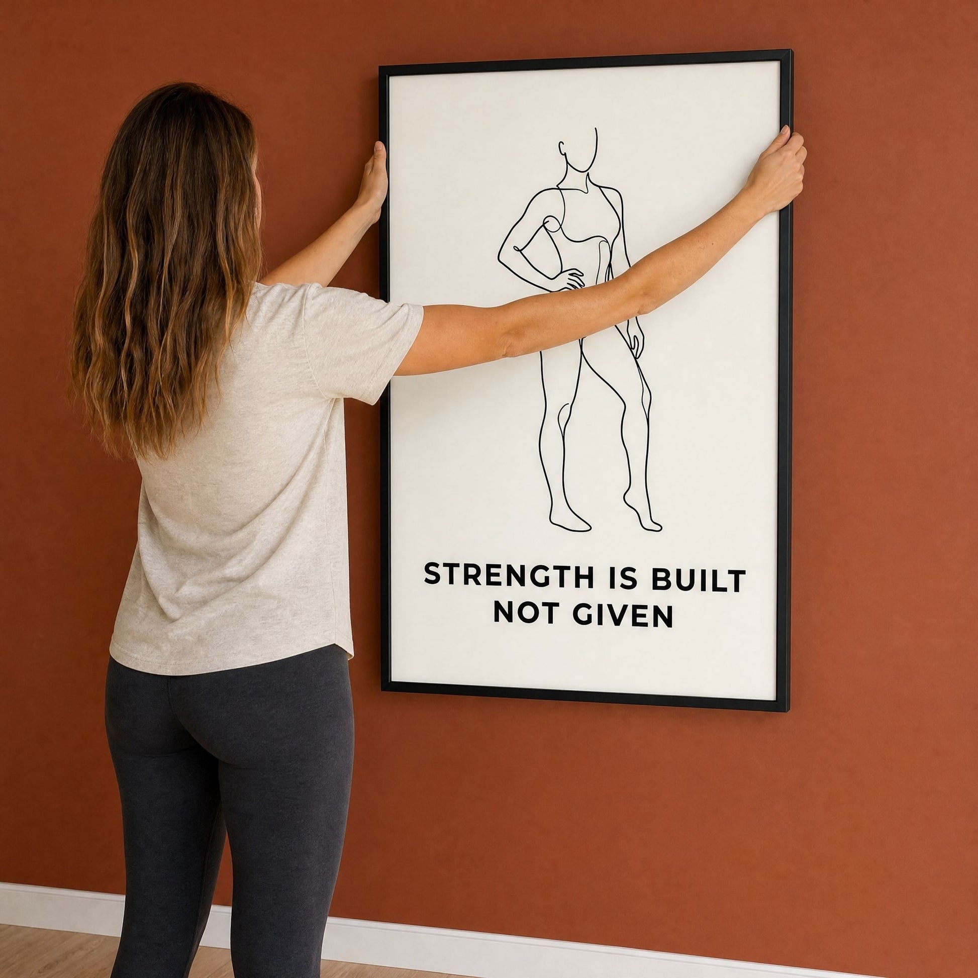 A woman reaches up to hang the black framed print on a terracotta wall, seen from behind