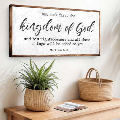 White canvas sign with black script, But seek first the kingdom of God, Matthew 6:33, above a wood console and a potted plant