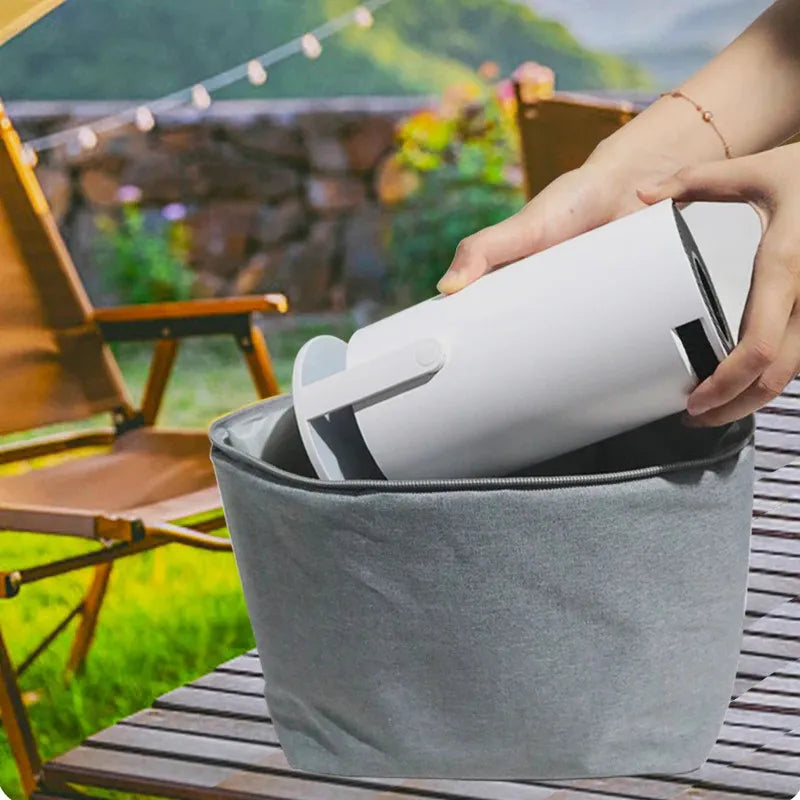 Person placing a white mini projector into a gray bag outdoors on a wooden table with a garden background.