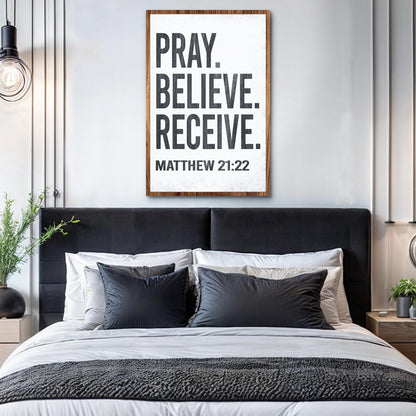 Tall white canvas sign with bold black block letters, Pray. Believe. Receive. Matthew 21:22, above a bed with black headboard