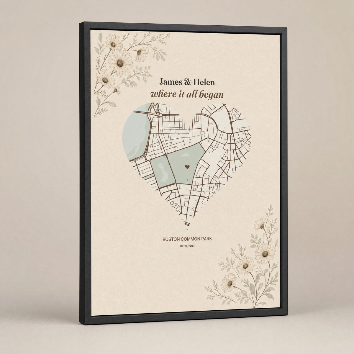 A heart-shaped city map print in a deep black frame, standing at a slight angle on a plain warm grey background, the names above the map and the place name and date beneath