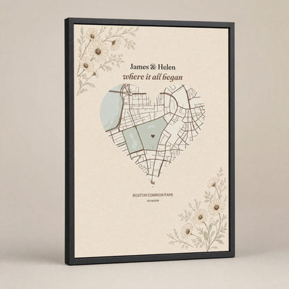 A heart-shaped city map print in a deep black frame, standing at a slight angle on a plain warm grey background, the names above the map and the place name and date beneath