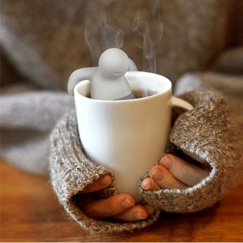 Person holding a steaming cup with a tea infuser shaped like a person.