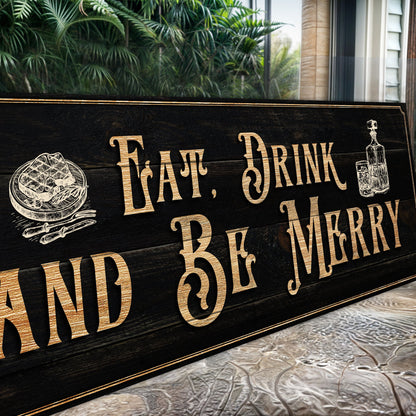 Angled close-up of the Eat, Drink and Be Merry canvas sign leaning outdoors on a patterned rug near tropical plants