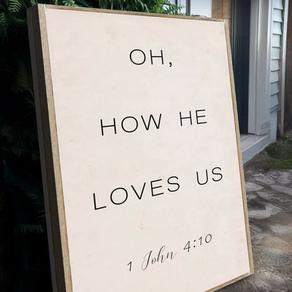 Angled close-up of the cream Oh How He Loves Us canvas sign with 1 John 4:10 in small script