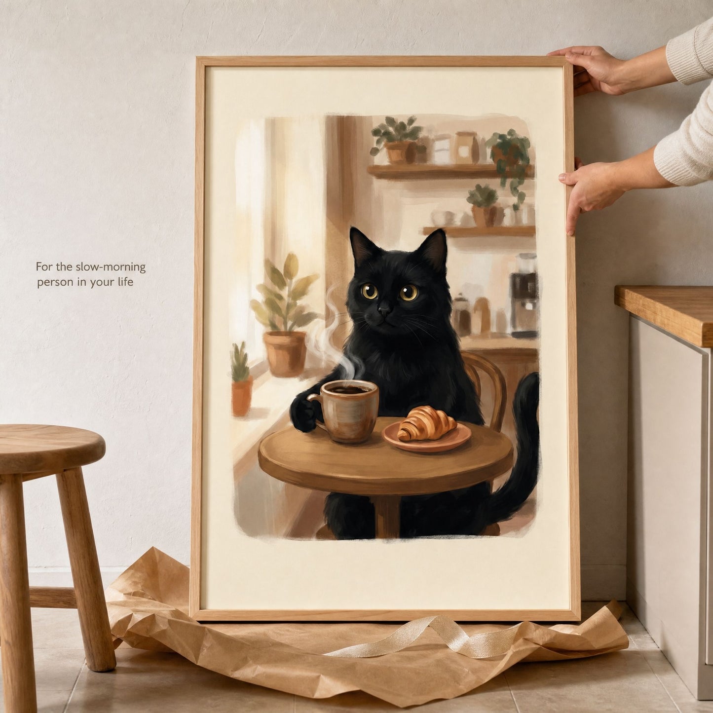 Hands lifting the framed black cat coffee print from its kraft paper wrapping beside a wooden stool