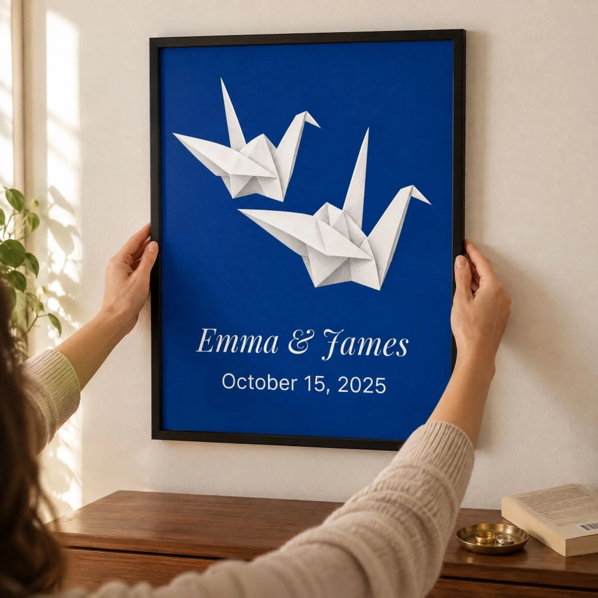 Hands levelling a framed paper cranes print on a bedroom wall in morning light