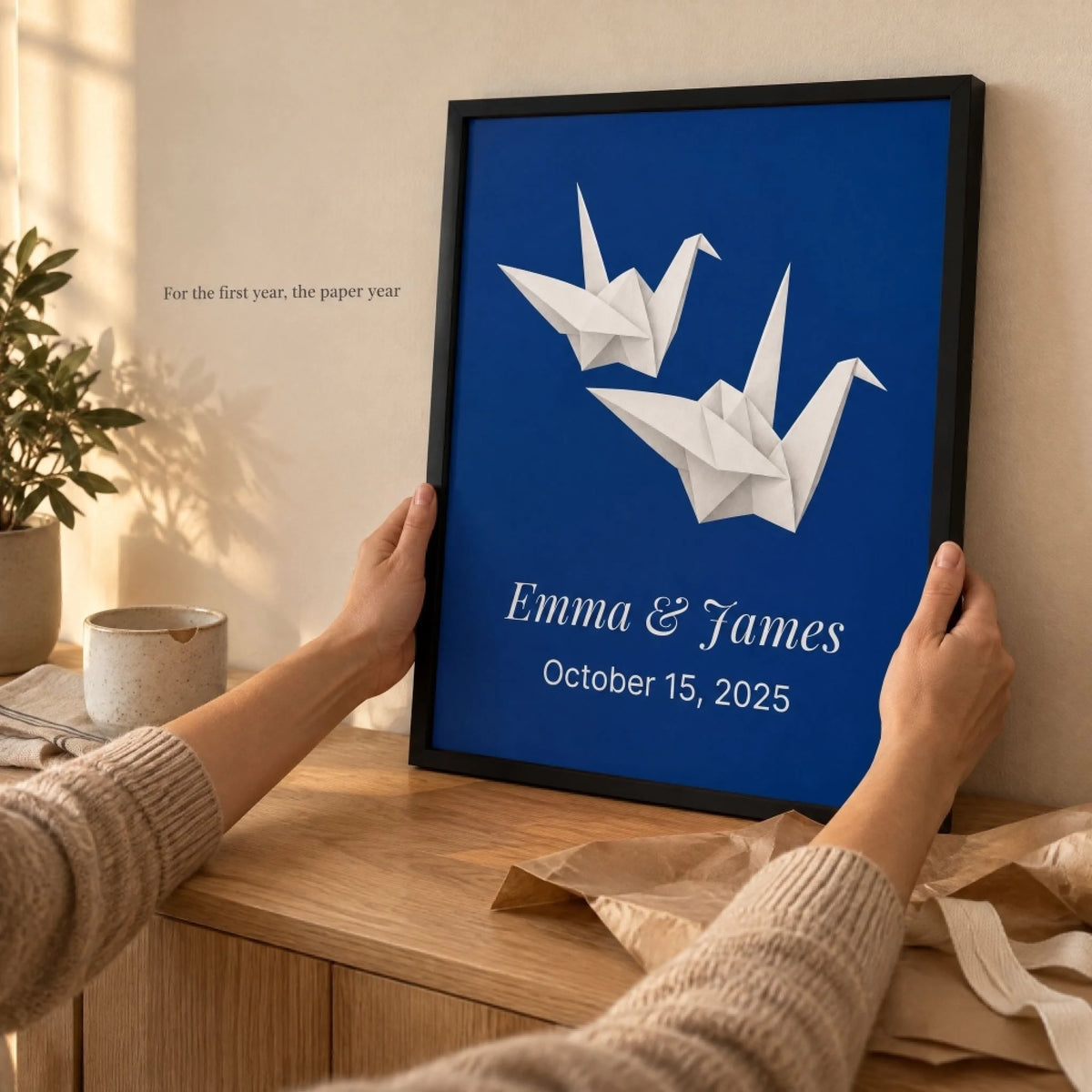 Hands holding the framed paper cranes anniversary print above a wooden table