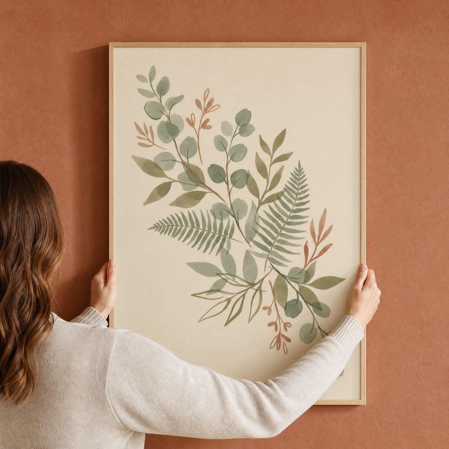 A woman hanging a large framed sage green eucalyptus and fern print on a terracotta wall