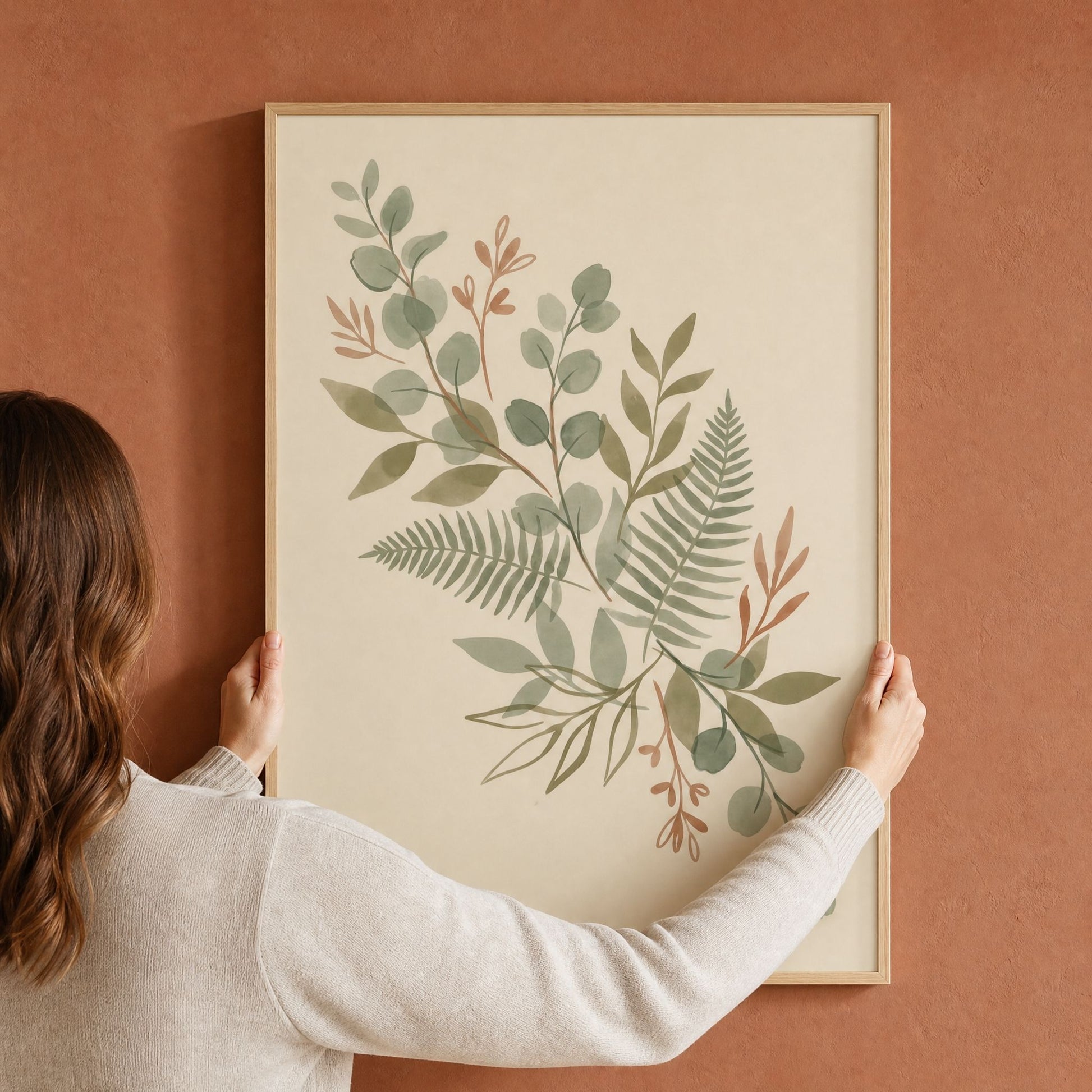 A woman hanging a large framed sage green eucalyptus and fern print on a terracotta wall