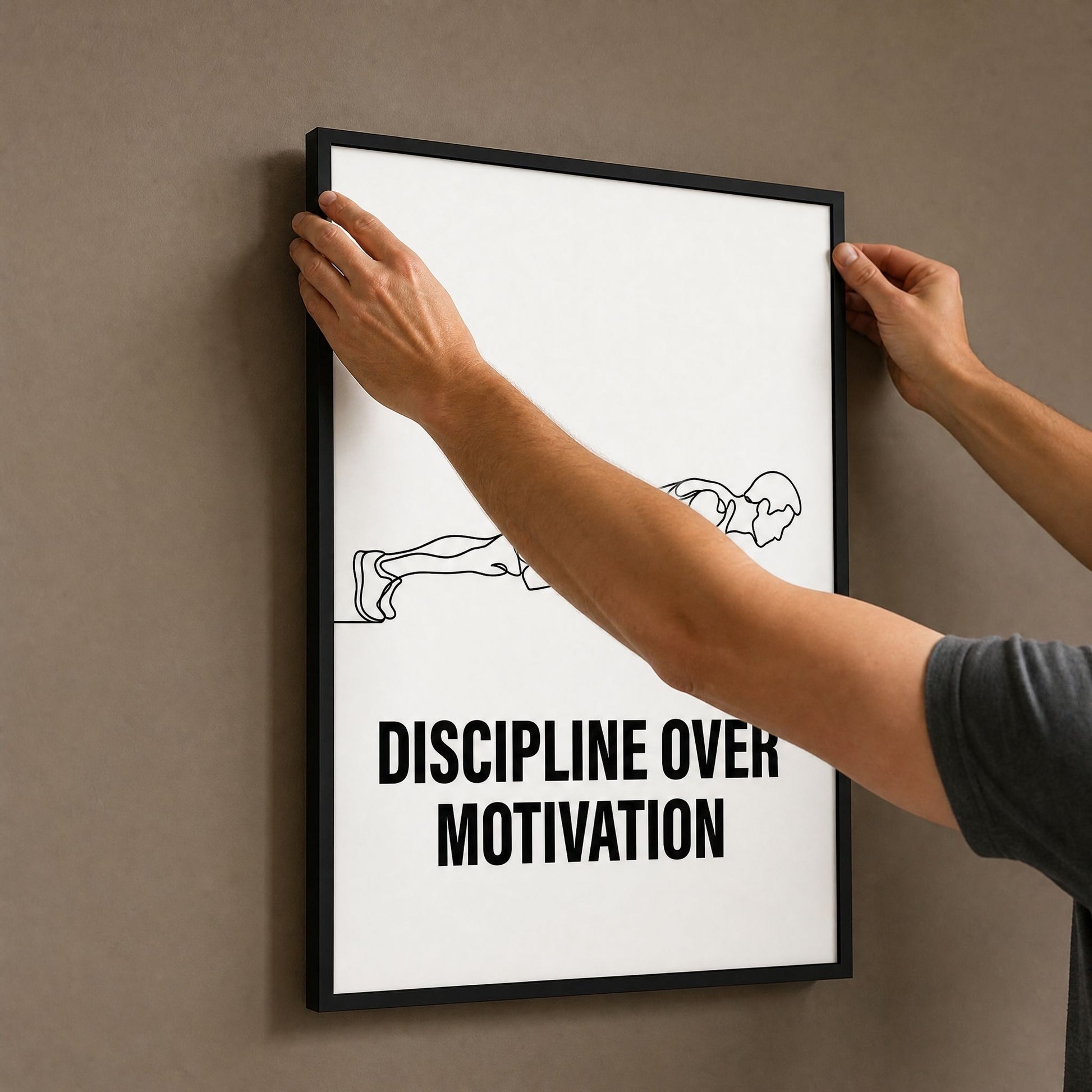 A person's hands hold the black framed print flat against a taupe wall while positioning it to hang