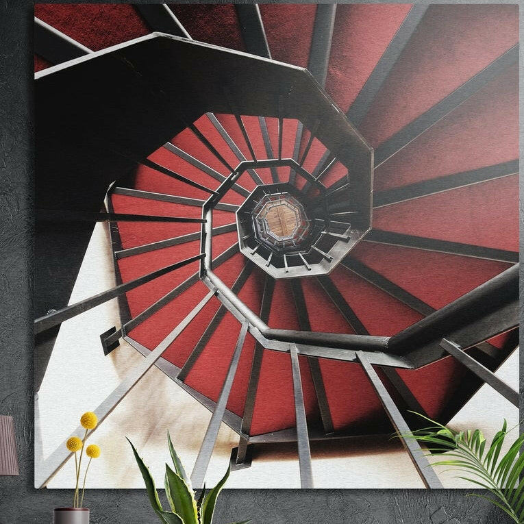Red spiral staircase seen from above with dark railings, brushed aluminum wall art on a dark wall behind plants