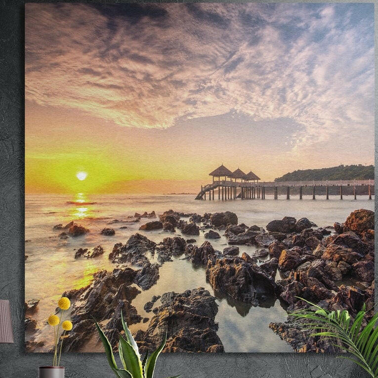Golden sunset over a rocky shore with a long pier and huts, brushed aluminum wall art on a dark wall behind plants