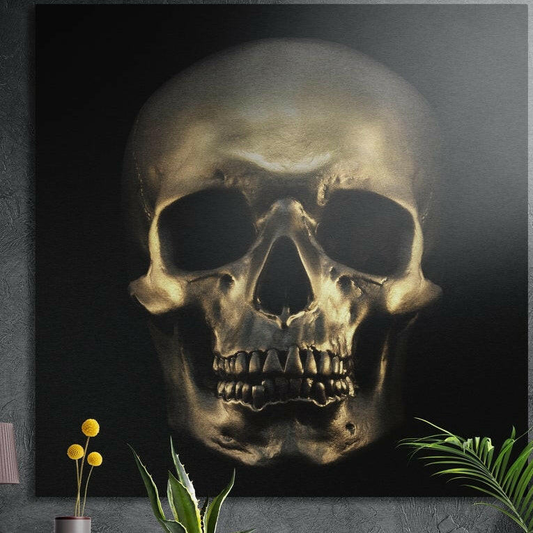 Polished bronze human skull facing forward against a deep black background, brushed aluminum wall art above potted plants