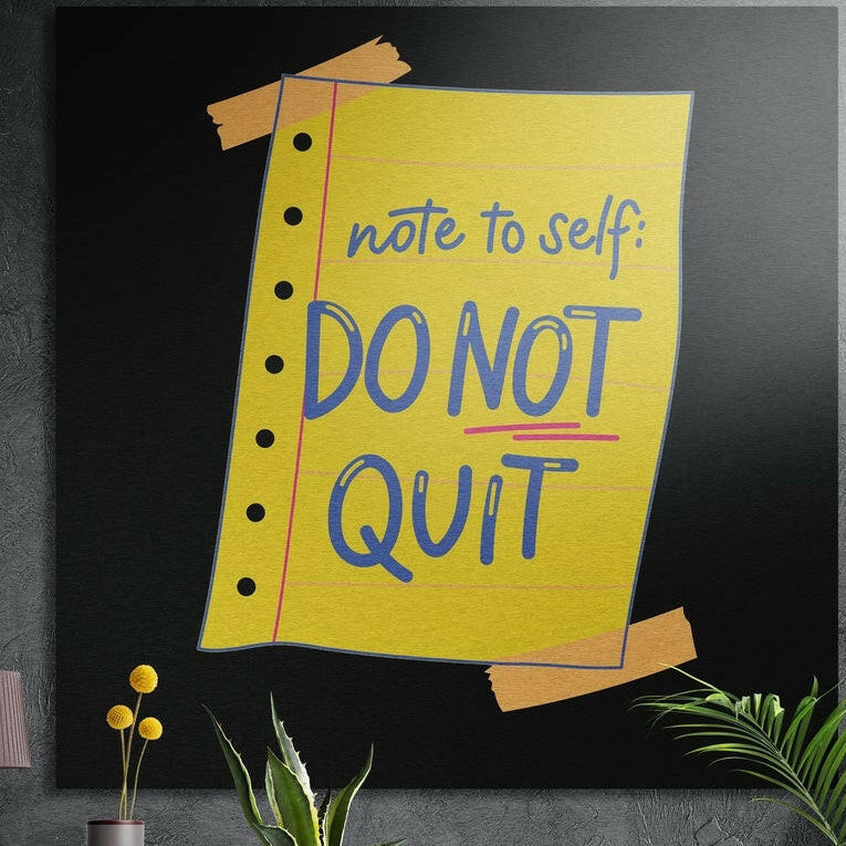 Taped yellow notebook note with blue motivational lettering on black, brushed aluminum wall art behind plants
