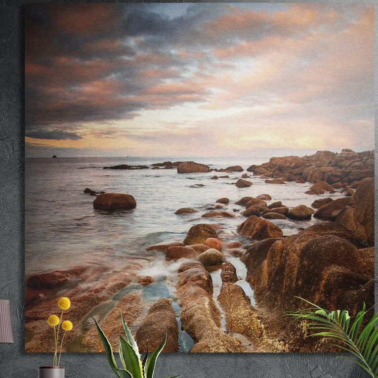 Rocky coast at sunset brushed aluminum wall art on a dark wall behind a vase of yellow flowers and plants