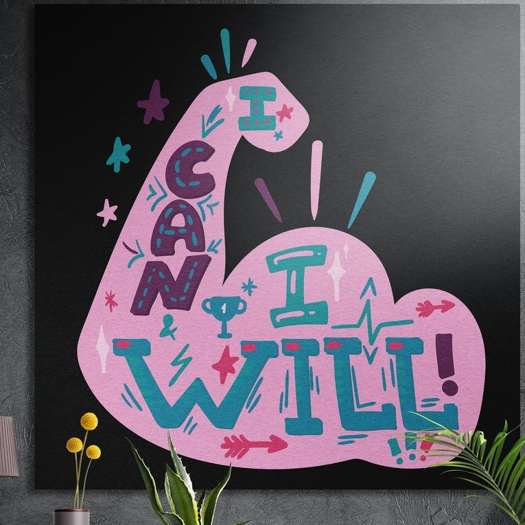 Pink flexed arm with I Can I Will lettering on black, brushed aluminum wall art on a dark concrete wall behind plants