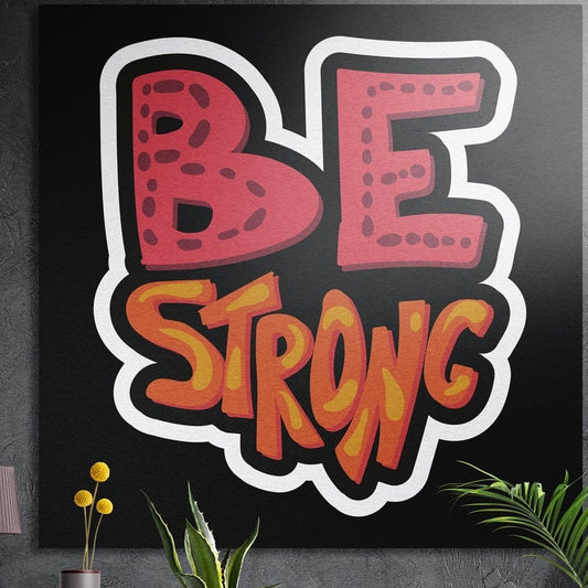 Be strong bold comic lettering brushed aluminum wall art on a dark wall above yellow flowers and plants