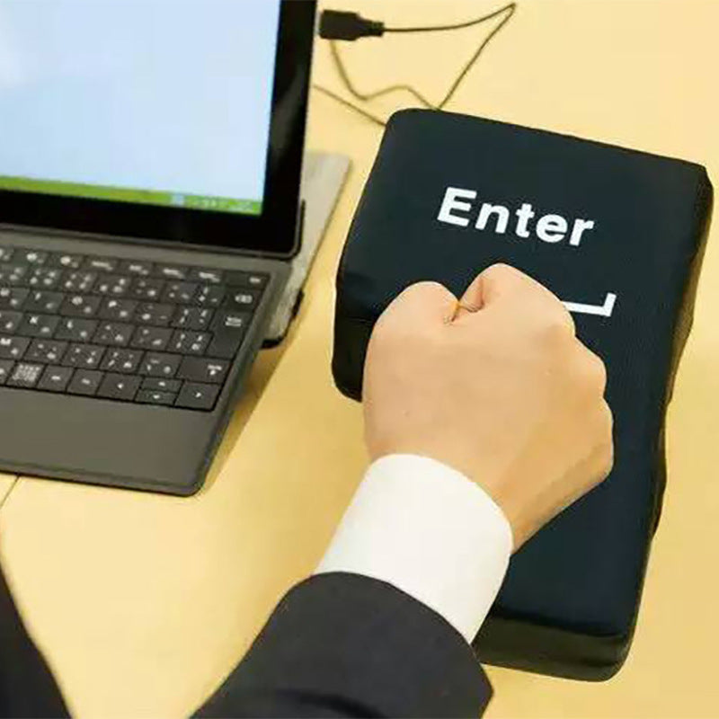 A person's hand punching a large, black, stress-relief USB Enter key on a desk, with a laptop and monitor in the background.
