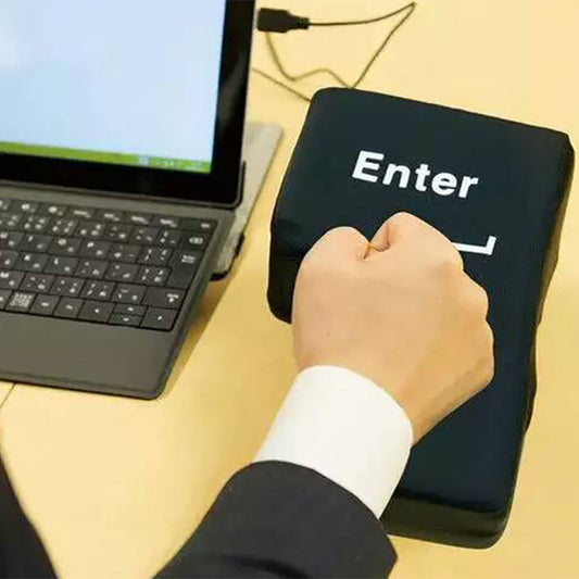 A person's hand punching a large, black, stress-relief USB Enter key on a desk, with a laptop and monitor in the background.