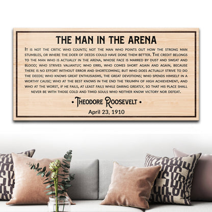 Light natural wood-look canvas sign with black Man in the Arena quote above a sofa with pink pillows