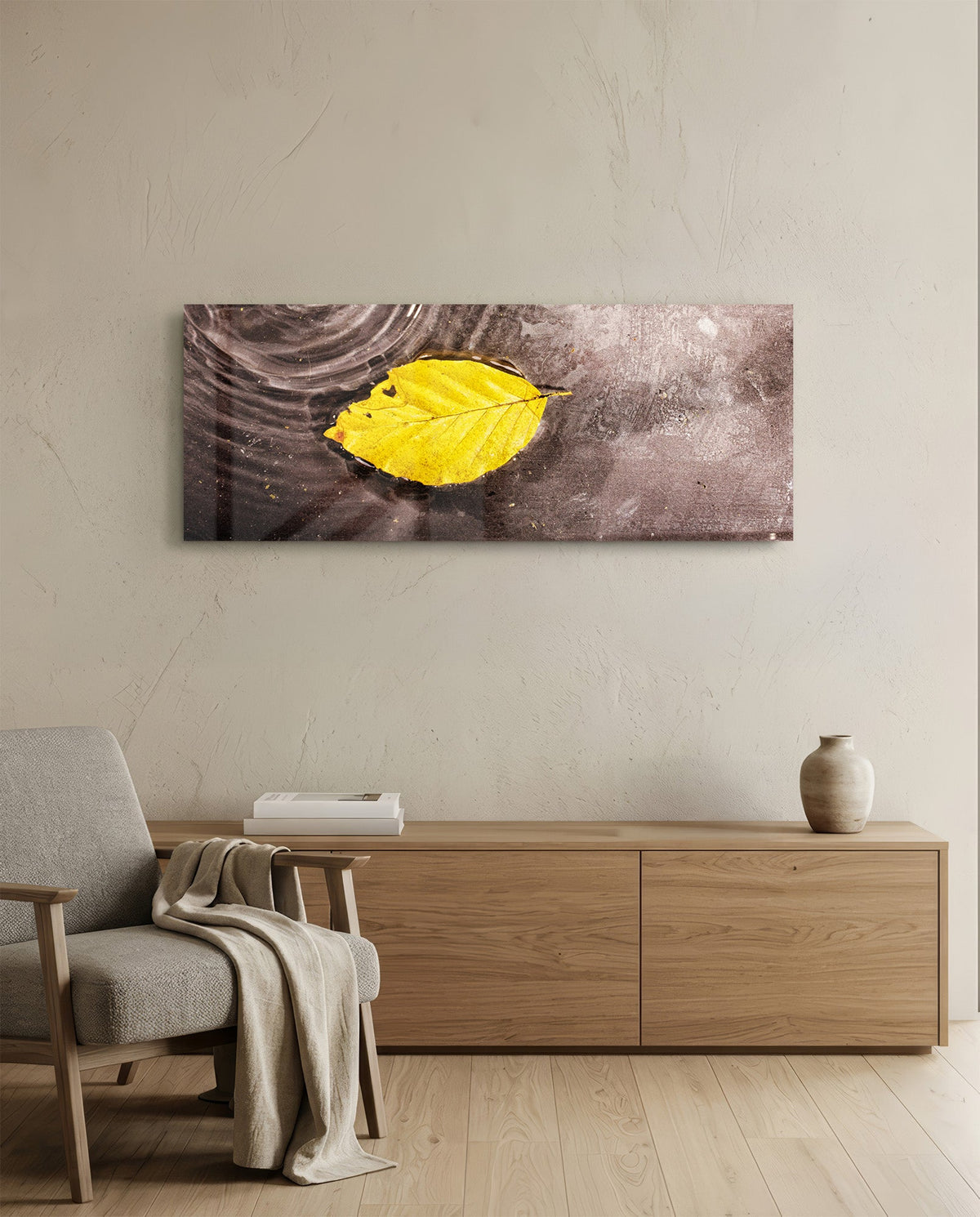 Yellow leaf on rippled water glass wall art above an oak sideboard beside a wooden armchair with a throw