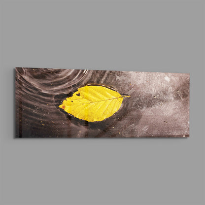 Yellow leaf on rippled water panoramic glass wall art shown on a plain gray background
